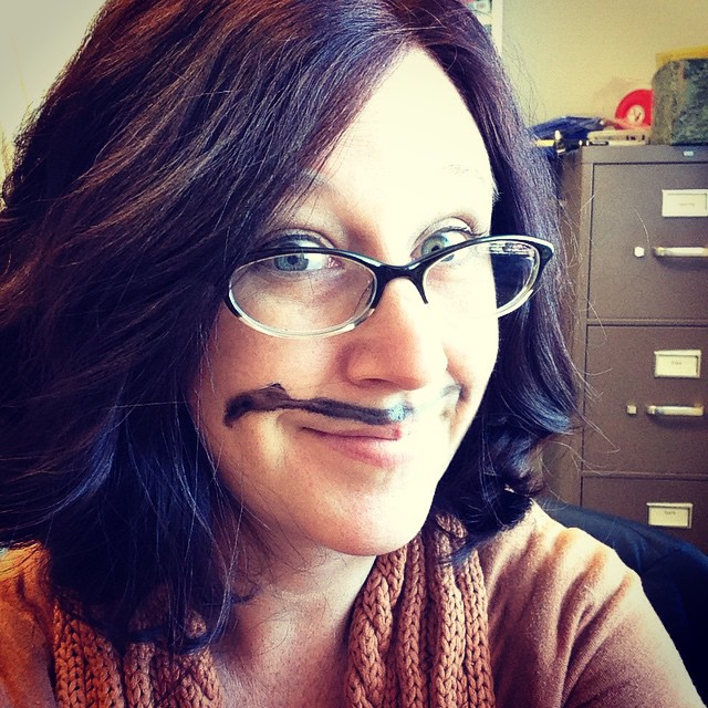 A caucasian woman with dark hair has a black mustache painted on her face.