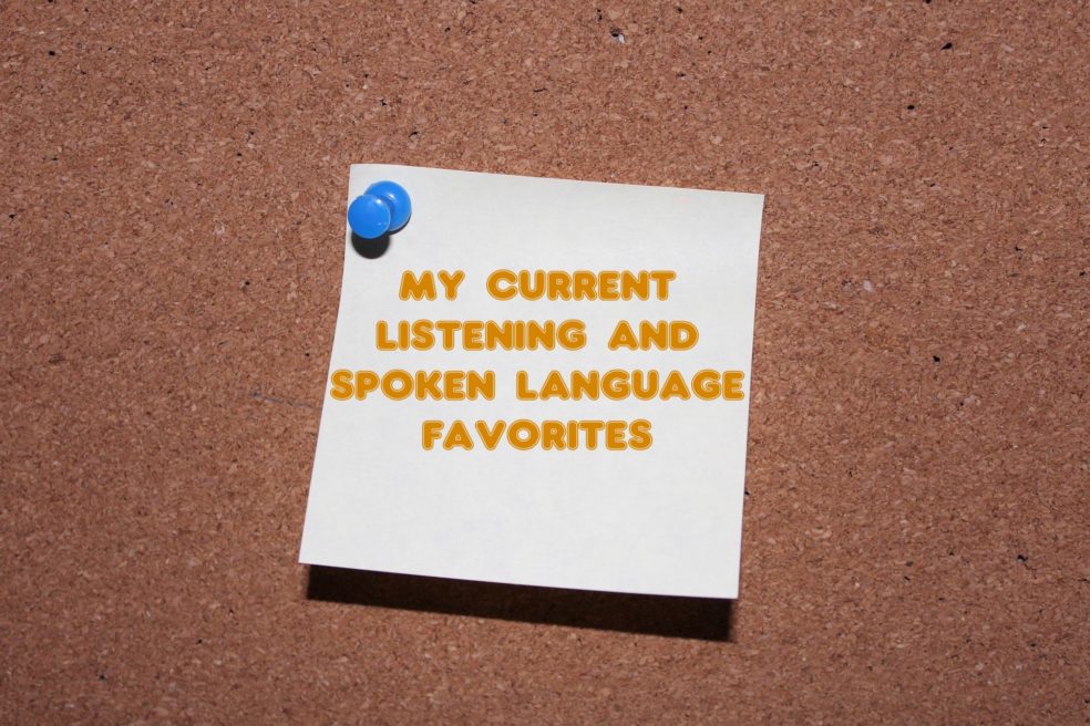 A white sticky note pinned to a cork board with the words "my current listening and spoken language favorites" in orange on the note