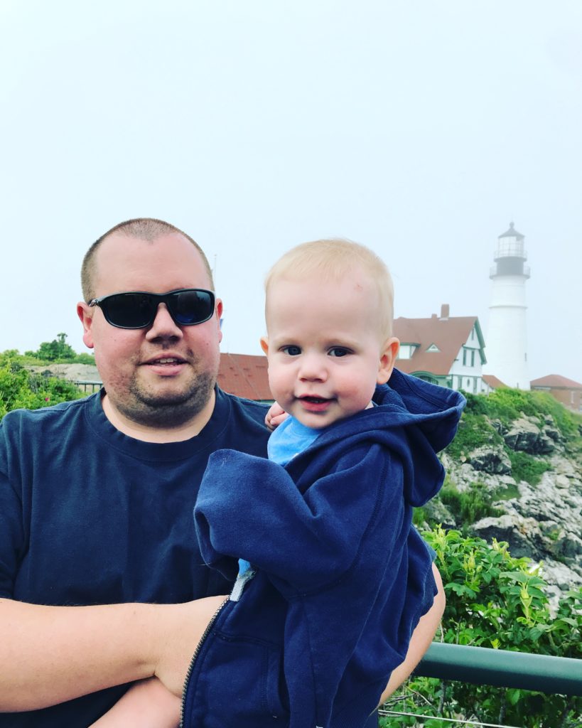 An image of a white man holding a white baby in front of a lighthouse.