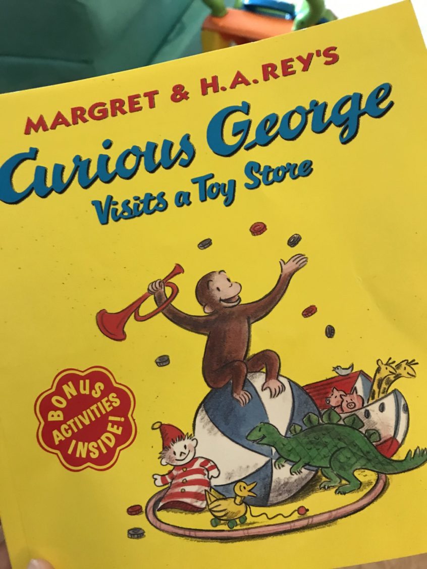 A picture of a yellow book with the title "Curious George Visits a Toy Store." There is a picture of a monkey sitting on a pile of toys.