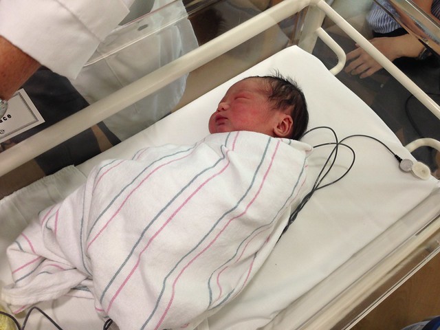 A newborn baby with dark hair laying in a bassinet in a newborn nursery.