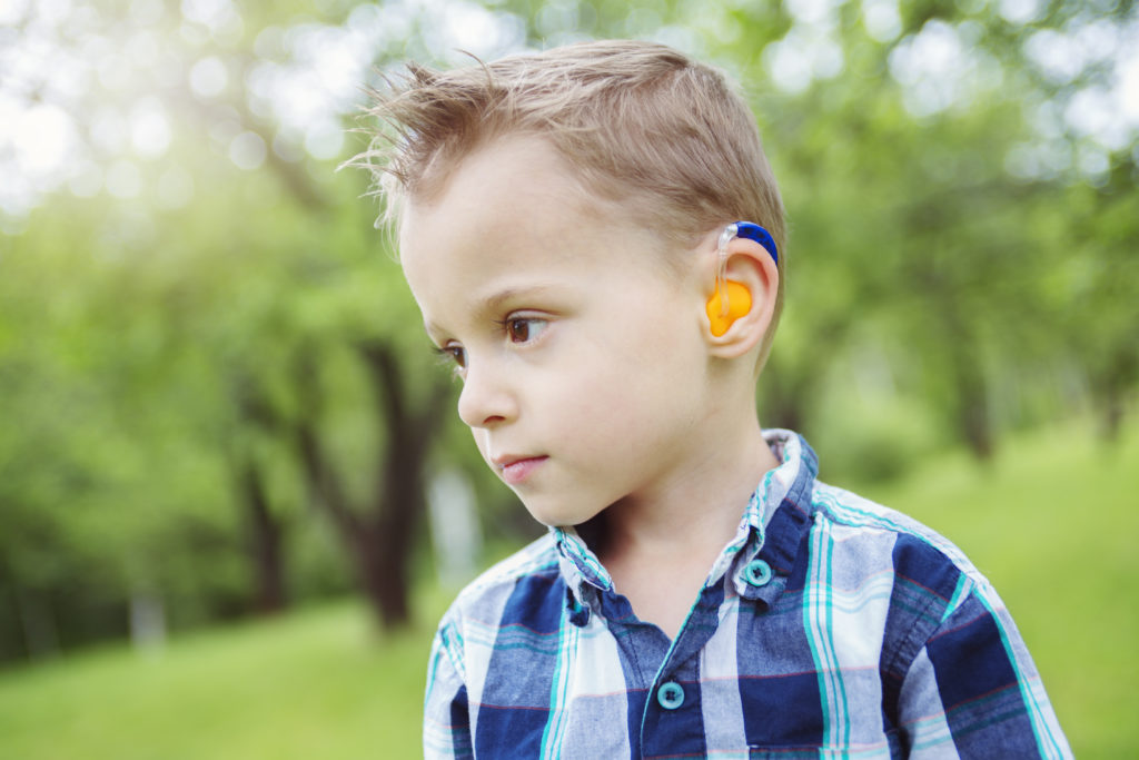 A caucasian boy with short hearing hearing a hearing aid with an orange earmold and blue ear piece.
