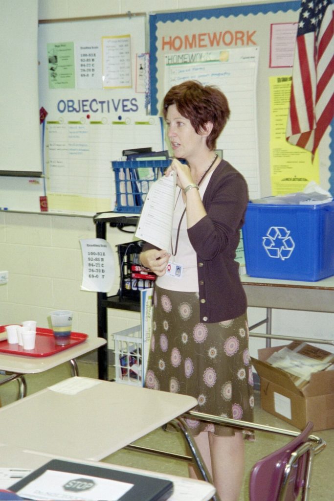 A photo of a Caucasian woman standing in the front of a classroom, holding up a sheet of paper. She is teaching, and has a bulletin board behind her. 
