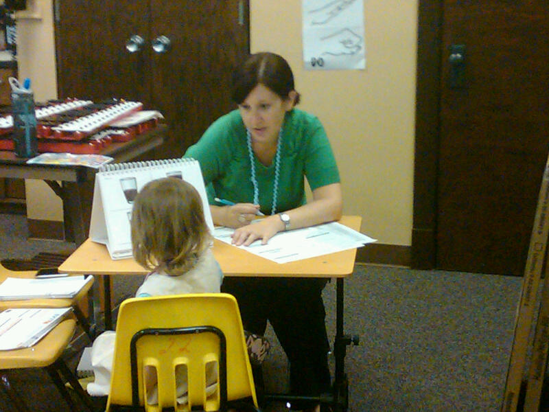 A caucasian woman wearing a green shirt is seated at a small table.  On the opposite side of the table the back of a child's head is visible. An easel with photos is visible on the table, and the woman is holding a pencil over a test protocol.