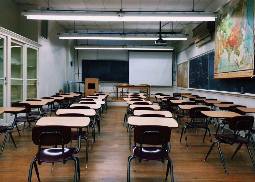 An image of a large classroom with desks in rows, a map on the wall and large flourescent lights hanging from the ceiling.