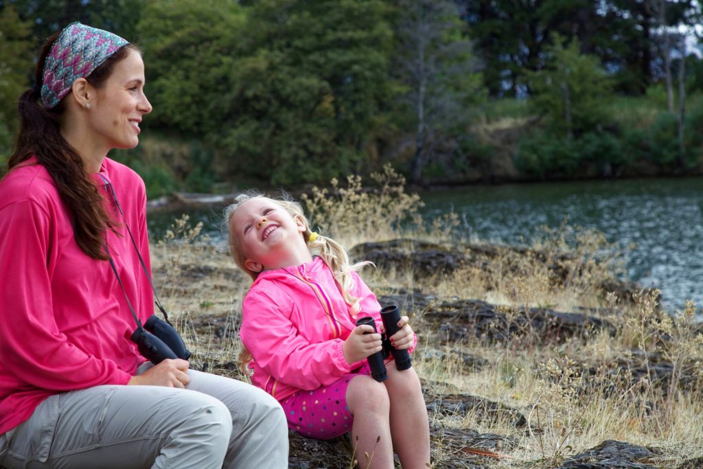 A white mother and daughter, both wearing pink jackets and holding binoculars, are seated on rocks near the edge of a body of water. The mother is smiling and the daughter is looking up at the mother laughing. 