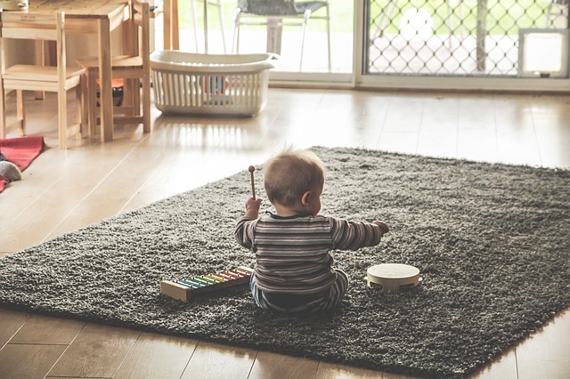 An image of a white baby, from the back, seated on a rug.  He has a xylophone on his left, and a tambourine on his right.  He is holding up a wooden mallet in his left hand.