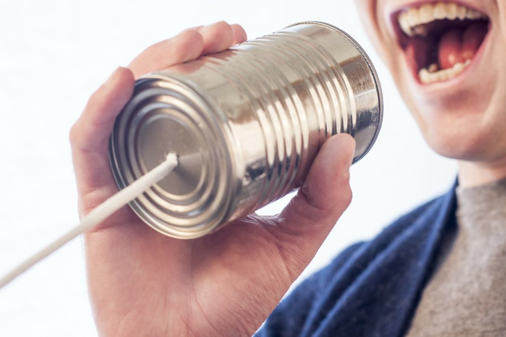 A person's mouth is visible. They are holding a tin can with a string attached to the end and talking into the can.