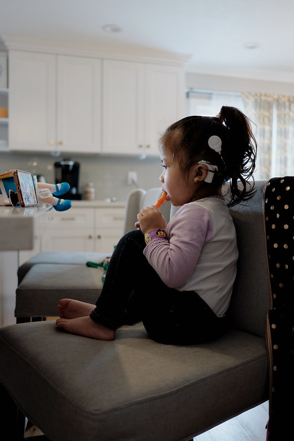 A girl is sitting on a chair watching a screen while holding a carrot. She has a white cochlear implant on her head. 