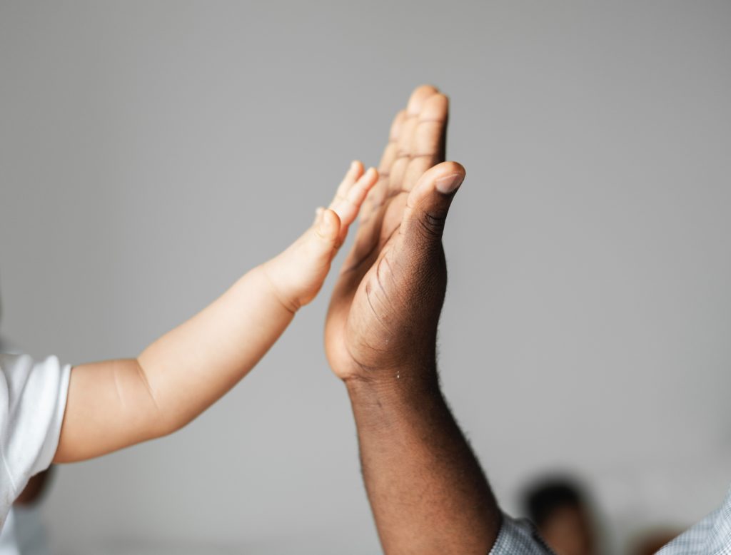 A baby's hand and an adult hand are both visible, the baby and adult appear to be high-fiving.
