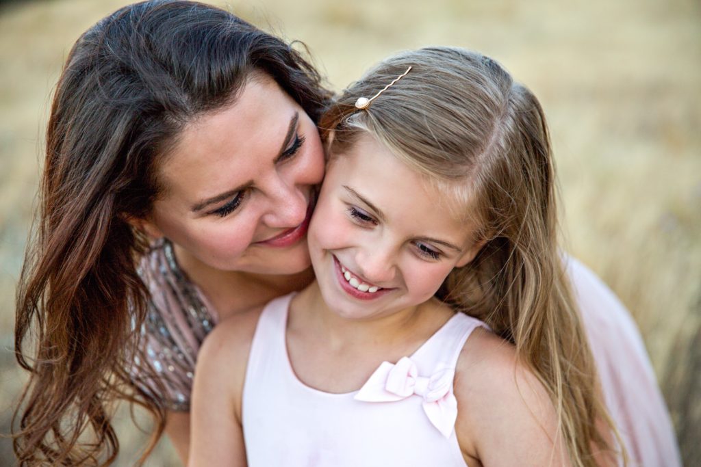 A mom is standing behind her daughter, hugging her from behind