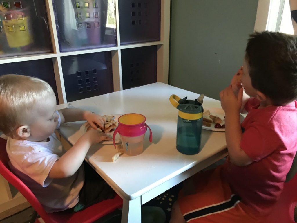 Two children sitting at a small table eating sandwiches