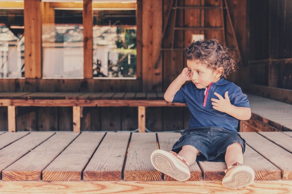 An image of a child sitting on a deck, pointing to his ear and listening. 