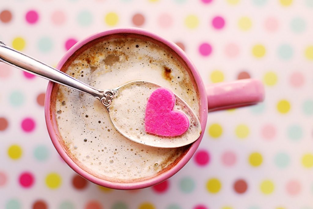 A mug of hot cocoa from above, set on a polka dot background. On the top of the hot cocoa is a spoon with a pink heart on it. 