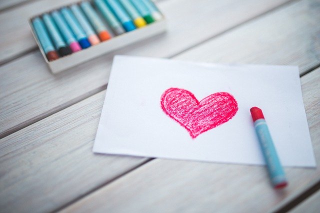 A white paper is on a table with a pink heart colored on it. There is a pink crayon out and a set of crayons on the table.